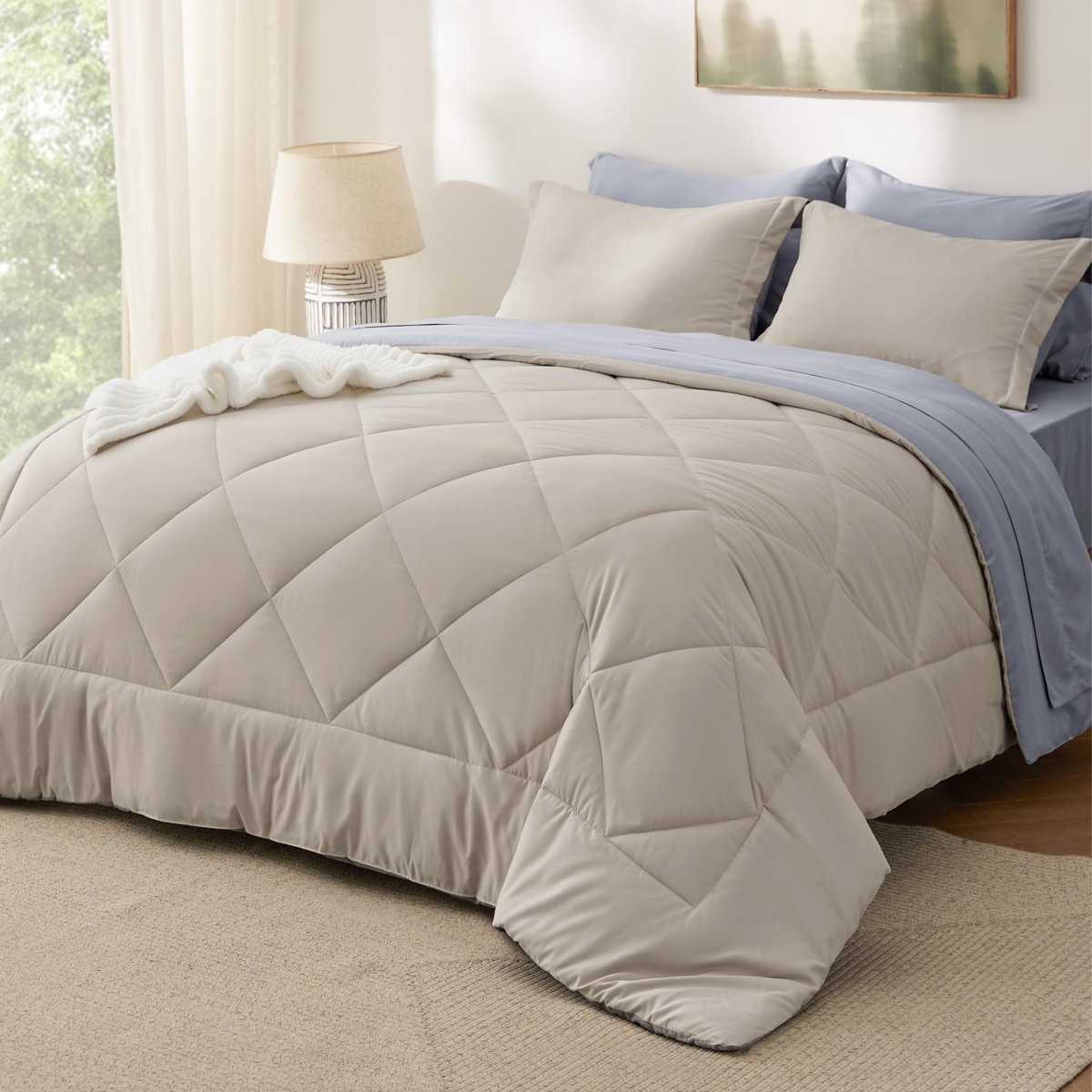 A neatly made bed features a beige quilted comforter with matching pillowcases and light blue sheets. A striped lamp and a framed picture are visible in the background.