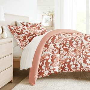 A bed features a floral-patterned comforter and matching shams in shades of peach and white. The room has a light wood nightstand and a vase with greenery.