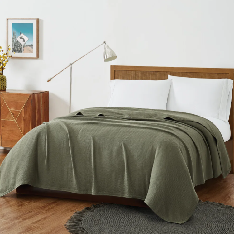 A neatly made bed features an olive green blanket and white pillows, with a wooden headboard. The room includes a wooden side cabinet, a modern floor lamp, a decorative plant, and a circular gray rug on wood flooring.