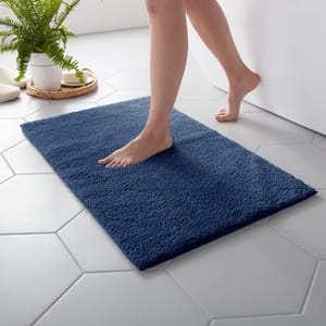 A person steps onto a blue bath mat placed on a hexagonal-tiled floor beside a white bathtub, with a potted fern and slippers in the background.