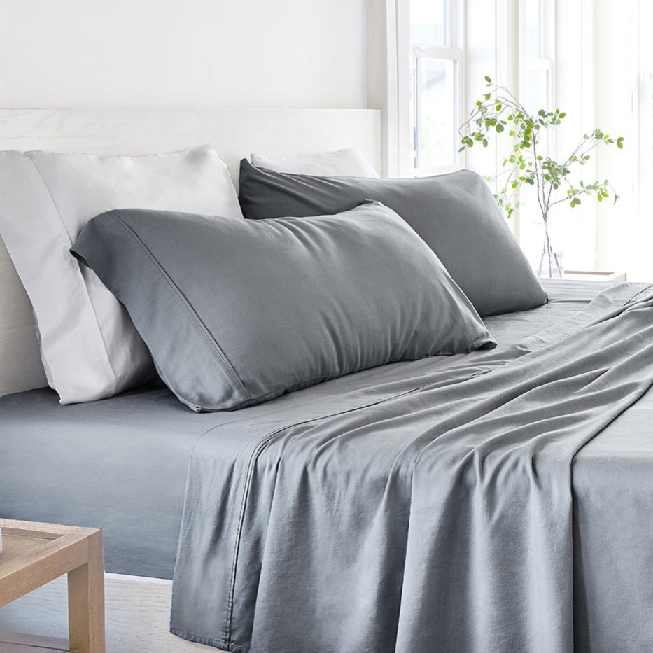 A bed set in gray tones features a fitted sheet, flat sheet, and two pillowcases, placed in a bright room with a plant nearby.