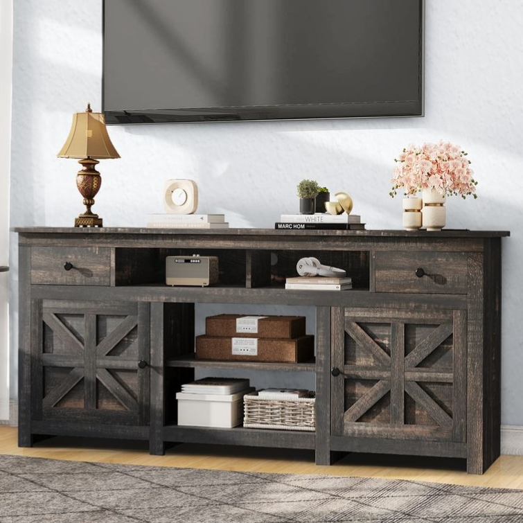 Rustic TV stand with a dark wood finish featuring two drawers, two closed cabinets with X-patterned doors, and open shelving. Decor includes a table lamp, books, a small plant, and decorative boxes.