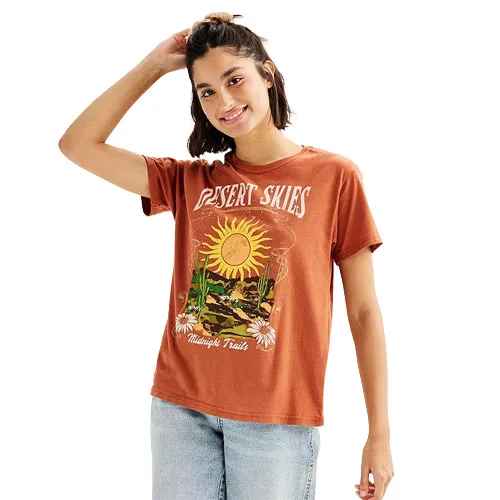 A woman wears a rust-colored t-shirt featuring a \“Desert Skies\“ graphic with a sun, cacti, and daisies, paired with light blue jeans.
