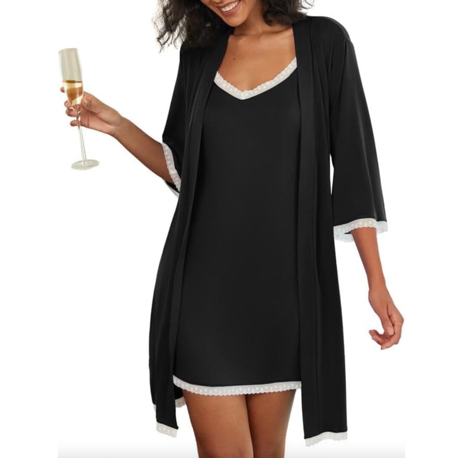 A woman wearing a black robe with white trim, holding a champagne glass. The robe features loose sleeves and falls above the knee, with matching trim on the dress underneath.