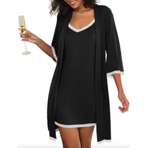 A woman wearing a black robe with white trim, holding a champagne glass. The robe features loose sleeves and falls above the knee, with matching trim on the dress underneath.