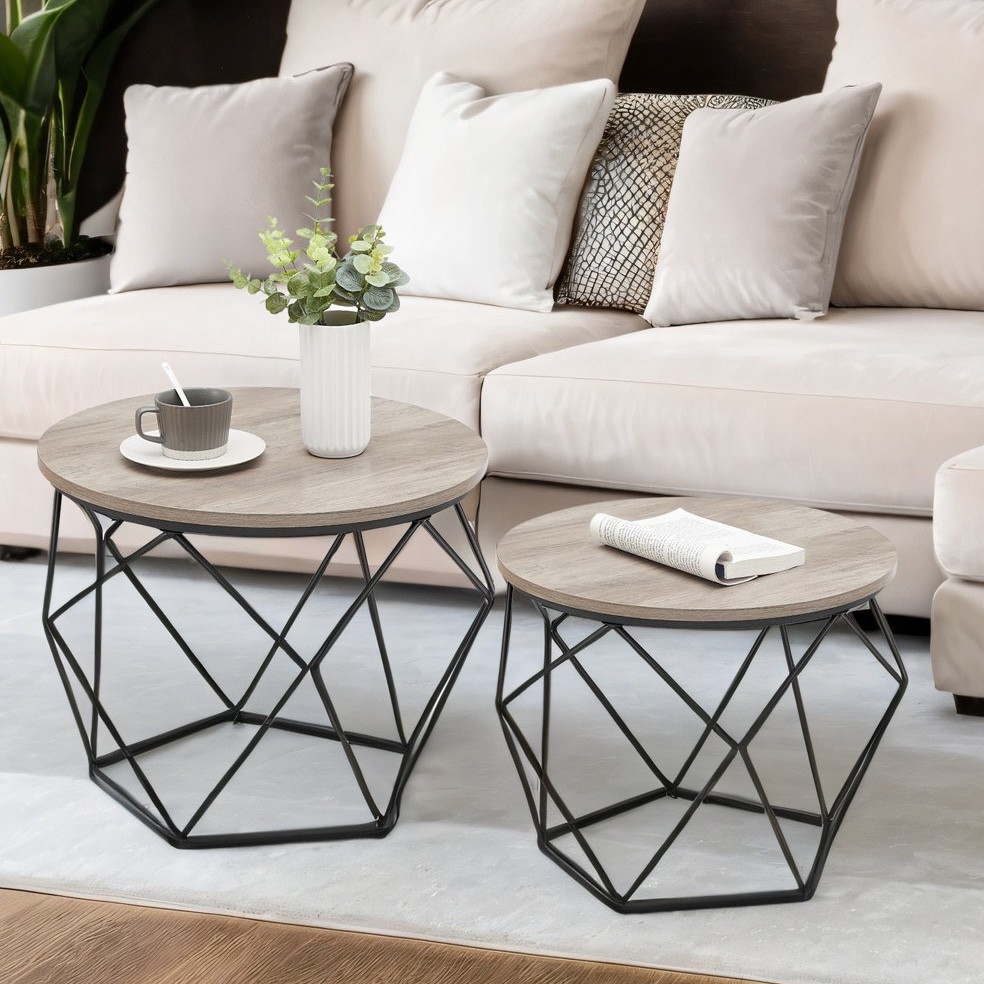 Two round coffee tables with wooden tops and geometric black metal bases, one larger and one smaller, are placed in front of a beige sofa. The larger table has a cup and saucer with a spoon and a potted plant, while the smaller table holds an open book.