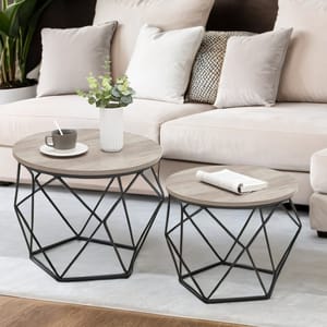 Two round coffee tables with wooden tops and geometric black metal bases, one larger and one smaller, are placed in front of a beige sofa. The larger table has a cup and saucer with a spoon and a potted plant, while the smaller table holds an open book.