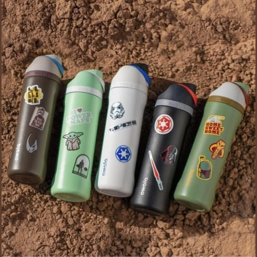 Five colorful Owala water bottles, each adorned with different themed stickers, are placed on a dirt surface.