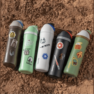 Five colorful Owala water bottles, each adorned with different themed stickers, are placed on a dirt surface.
