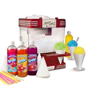 A snow cone machine with crushed ice inside, surrounded by Kool-Aid snow cone syrups in Sharkleberry, Peach Mango, and Black Cherry flavors, alongside colorful snow cones and accessories like plastic spoons and paper cups.