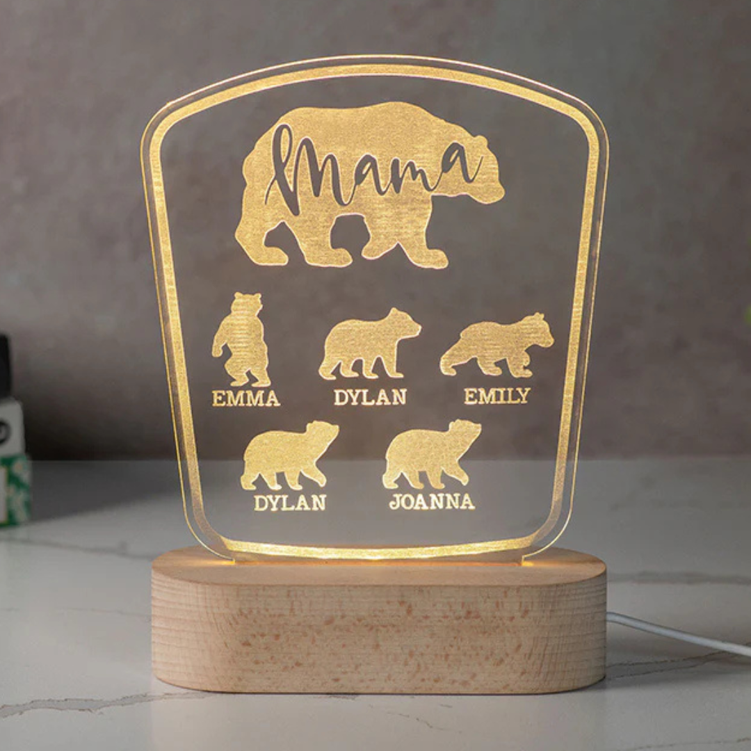 A glowing acrylic sign features a bear design personalized with the word \“Mama\“ and names Emma, Dylan, Emily, Joanna. The sign sits on a wooden base and is illuminated from below.