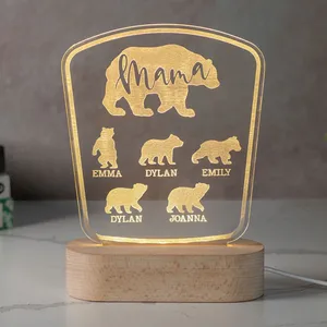 A glowing acrylic sign features a bear design personalized with the word \“Mama\“ and names Emma, Dylan, Emily, Joanna. The sign sits on a wooden base and is illuminated from below.