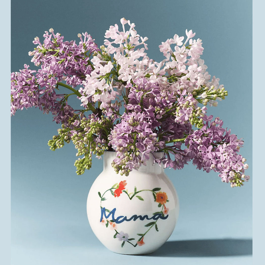 A vase with \“Mama\“ written on it, decorated with floral designs, holds a bouquet of lilacs in shades of light purple and pink against a blue background.