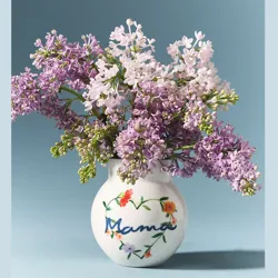 A vase with \“Mama\“ written on it, decorated with floral designs, holds a bouquet of lilacs in shades of light purple and pink against a blue background.