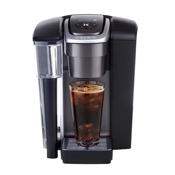 A Keurig coffee maker with a black finish, featuring a transparent water reservoir on the side, is brewing a tall glass of iced coffee placed on its drip tray.