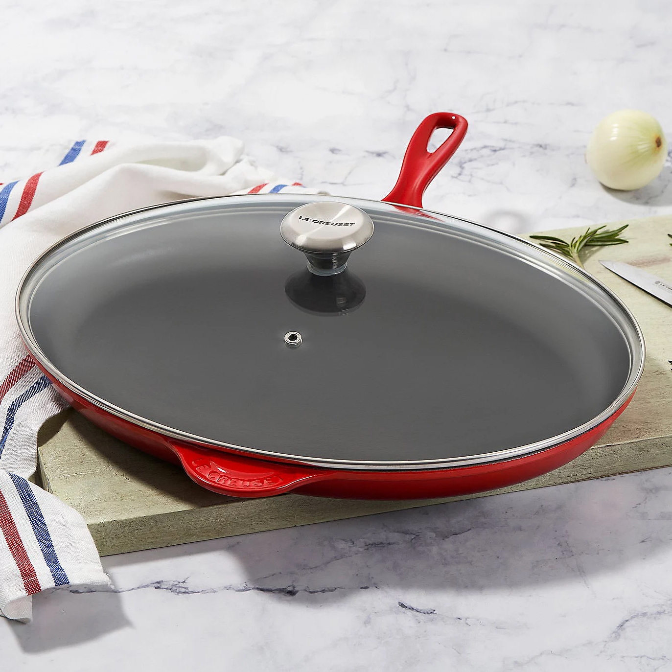Le Creuset Classic Oval Skillet is a red enameled cast iron pan with a matching glass lid and stainless steel knob. It features dual handles and is designed for even heat distribution and retention.