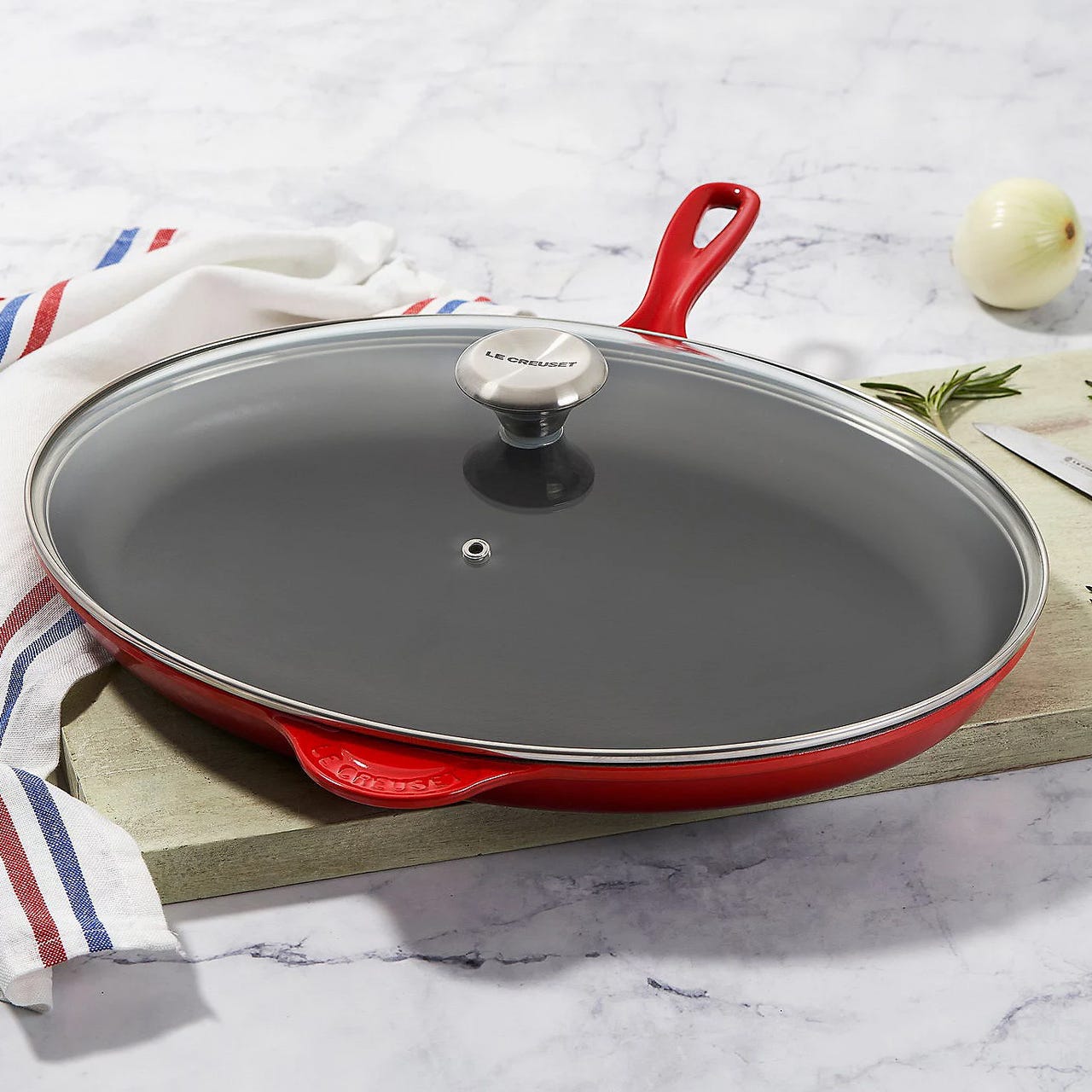 Le Creuset Classic Oval Skillet is a red enameled cast iron pan with a matching glass lid and stainless steel knob. It features dual handles and is designed for even heat distribution and retention.