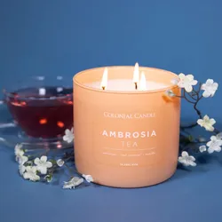 A Colonial Candle labeled \“Ambrosia Tea\“ with scents of nectarine, red currant, and vanilla is burning alongside a cup of red tea and decorated with small white flowers.