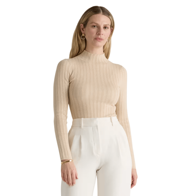 A woman is wearing a beige ribbed long-sleeve top with a high neck, paired with high-waisted white trousers. She accessorizes with gold hoop earrings and a bracelet.