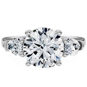 A three-stone engagement ring featuring a large round center diamond flanked by two smaller round diamonds, set in a white metal band.