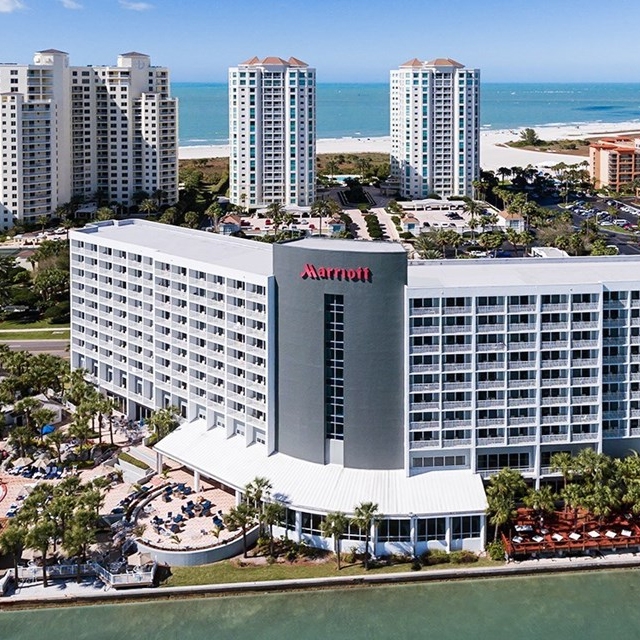 A multi-story Marriott hotel by the waterfront with surrounding palm trees and nearby high-rise buildings. The ocean is in the background.