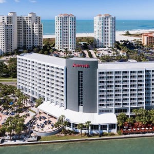 A multi-story Marriott hotel by the waterfront with surrounding palm trees and nearby high-rise buildings. The ocean is in the background.
