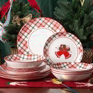 Festive dinnerware set featuring plates and bowls adorned with red and green plaid patterns and Christmas bell designs, set on a holiday-themed table with decorative fir branches and ribbon accents.