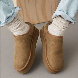 A pair of tan suede slip-on shoes worn with cream socks and blue jeans.