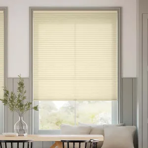 A beige cellular shade covers a window in a cozy, minimalist room. The shade is surrounded by a gray frame, with a wooden table and chairs, a glass vase with greenery, and a sofa with cushions nearby.