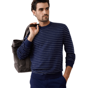 A man wearing a navy blue sweater with white stripes and holding a gray tote bag over his shoulder. He is also dressed in blue pants.