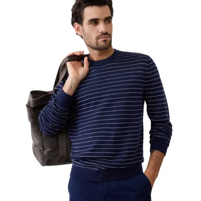 A man wearing a navy blue sweater with white stripes and holding a gray tote bag over his shoulder. He is also dressed in blue pants.