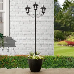 Tall planter with three black lantern-style lights, set against a white brick wall with a garden backdrop.
