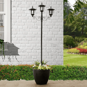 Tall planter with three black lantern-style lights, set against a white brick wall with a garden backdrop.