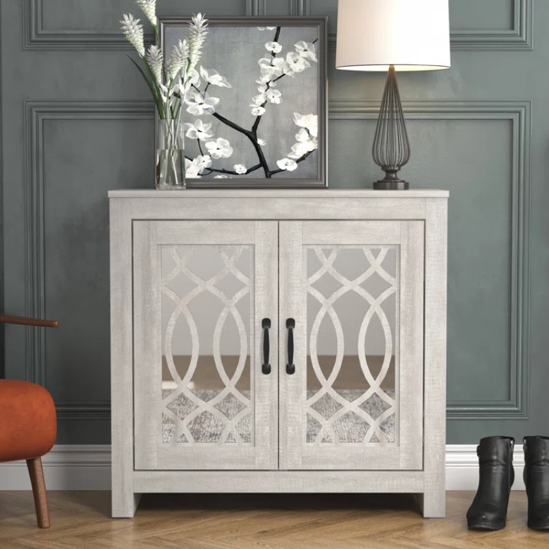 A decorative cabinet with mirrored doors featuring geometric patterns is topped with a floral painting, a vase with flowers, and a table lamp. An orange chair and a pair of black boots are nearby, adding to the room's decor.