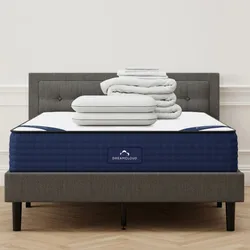 A grey upholstered bed with a blue DreamCloud mattress is topped with a stack of white pillows and folded bedding.