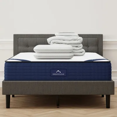 A grey upholstered bed with a blue DreamCloud mattress is topped with a stack of white pillows and folded bedding.