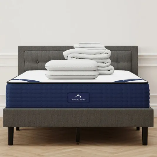 A grey upholstered bed with a blue DreamCloud mattress is topped with a stack of white pillows and folded bedding.