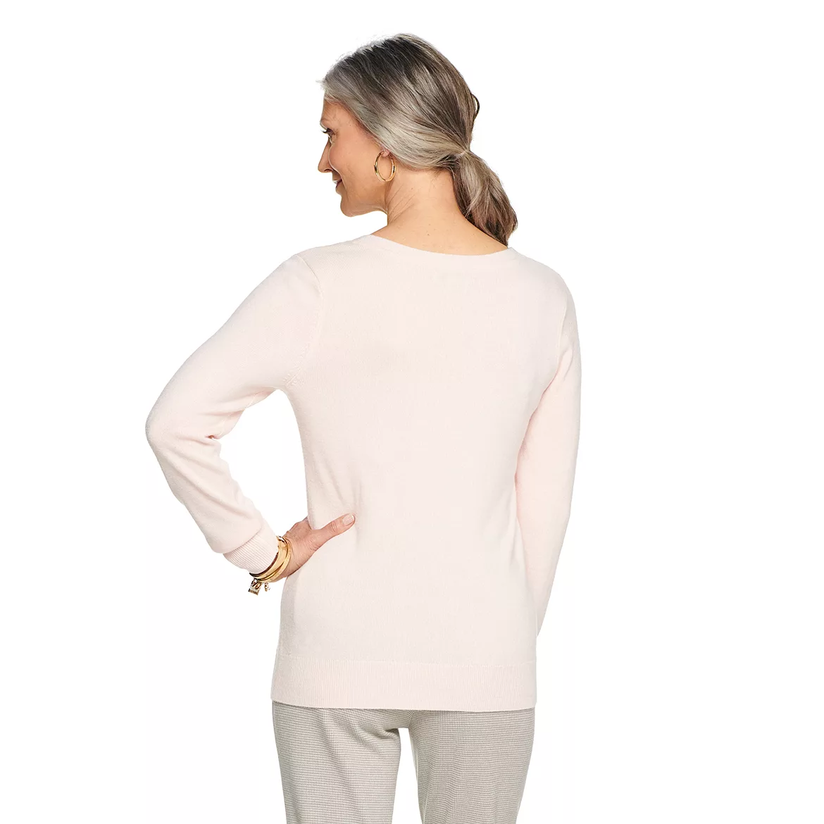 A woman is wearing a long-sleeve, V-neck sweater in a solid light pink color, with a relaxed fit that extends past the waistline.