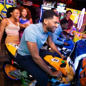 A group enjoys an arcade, focused on motorcycle racing games, with friends gathered around cheering.