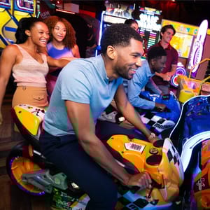 A group enjoys an arcade, focused on motorcycle racing games, with friends gathered around cheering.