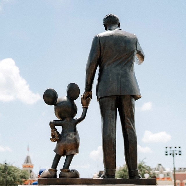 A statue of a man holding hands with Mickey Mouse, both facing away, under a clear blue sky.