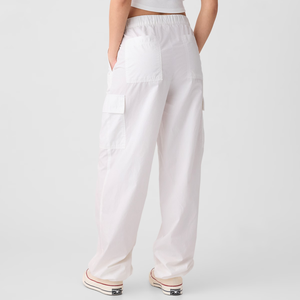 White parachute cargo pants with a high waist and elastic waistband, featuring large side pockets and a relaxed, wide-leg fit. Visible footwear includes pink and white sneakers with a black stripe.