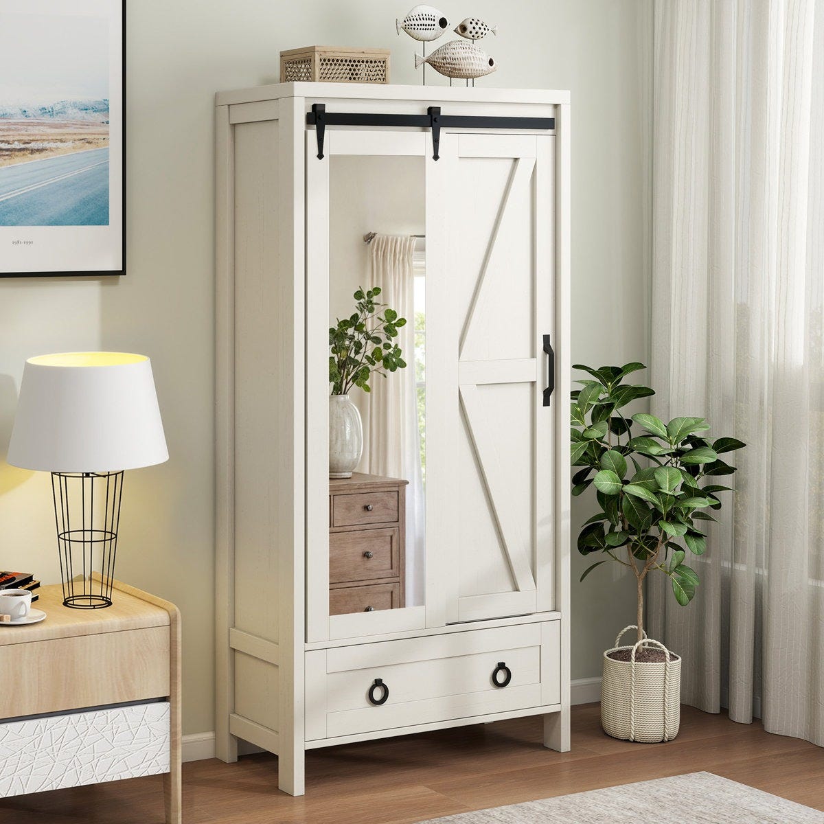 A white wardrobe with a sliding barn door, mirror, and bottom drawer sits beside a modern lamp on a small side table. A potted plant is on the floor nearby, and decorative items are placed atop the wardrobe.