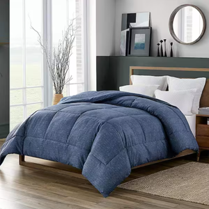A plush, blue, quilted comforter made with ultra-soft down alternative material covers a modern wooden bed, accompanied by white pillows. The room features a contemporary design with large windows, neutral walls, and minimalistic decor.