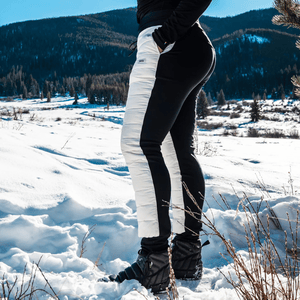 A person stands in the snow wearing black-and-white insulated pants and black puffer-style snow boots, set against a snowy mountain landscape.