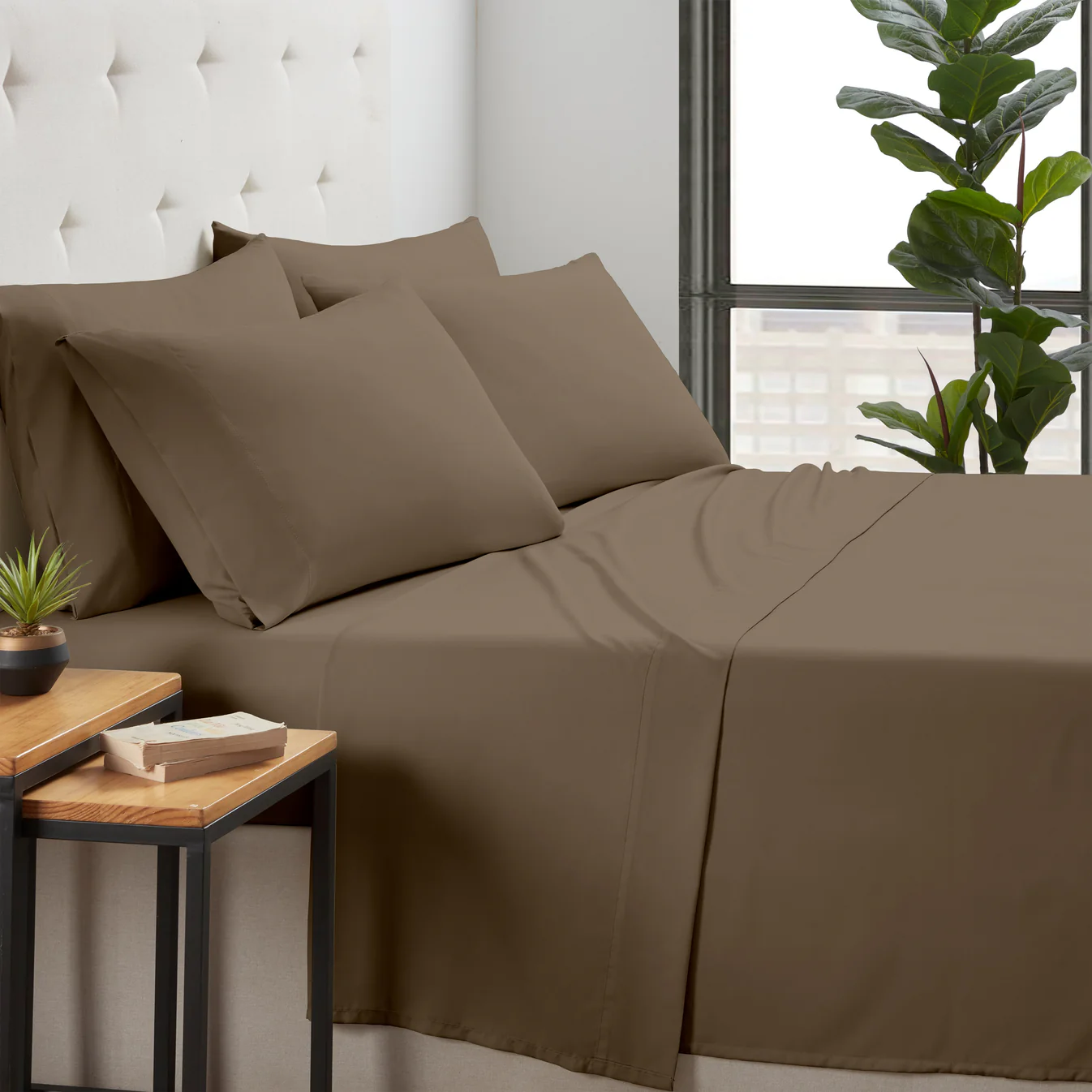 Brown bedding set with four pillows on a bed, accompanied by a small plant and a book on a wooden side table, near a large window.