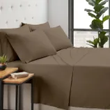 Brown bedding set with four pillows on a bed, accompanied by a small plant and a book on a wooden side table, near a large window.