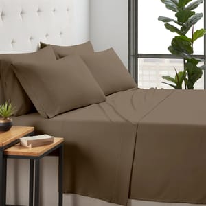 Brown bedding set with four pillows on a bed, accompanied by a small plant and a book on a wooden side table, near a large window.