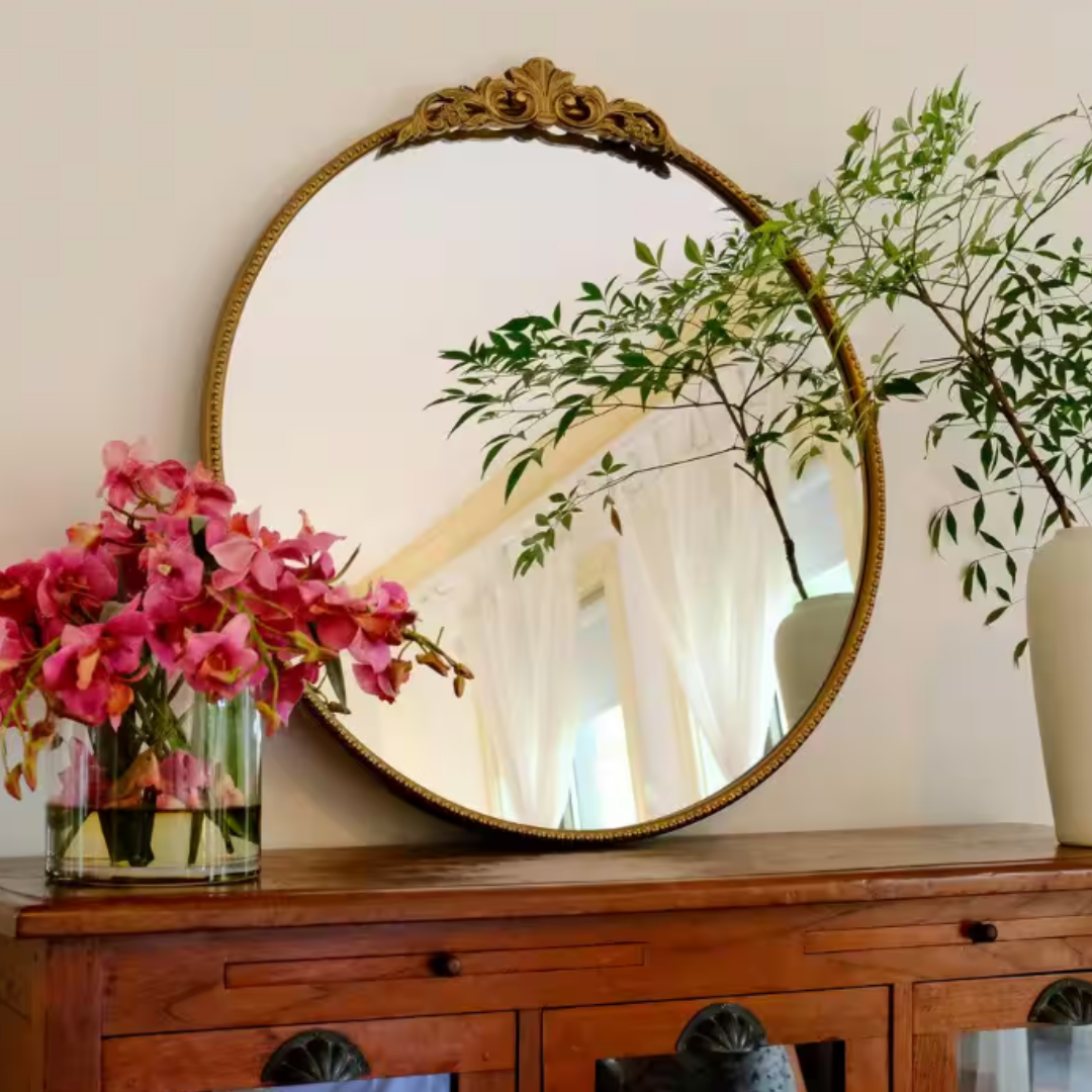 A round, ornately-framed mirror is mounted above a wooden cabinet adorned with a vase of pink flowers and another with green foliage.