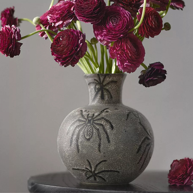 A gray vase with spider designs holds dark red ranunculus flowers.